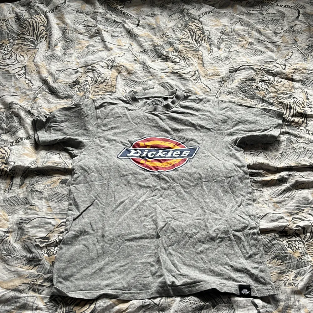 Säljer en grå t-shirt från Dickies med stor färgglad logga på bröstet. Klassisk passform och rund halsringning. Perfekt för en avslappnad stil och enkel att matcha med jeans eller shorts.. T-paidat.
