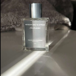 Aegean Salt & Citrus Eau de Parfum - Elegant parfymflaska med en fräsch doft av Aegean Salt & Citrus. Flaskan är fyrkantig och genomskinlig med en stilren etikett i silver och en matchande silverfärgad kork. Perfekt för den som älskar friska och marina dofter.
