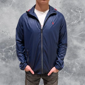 Ralph Lauren Windbreaker Jacka Blå - •Ralph Lauren Windbreaker jacka blå. Skick: 8/10, Inga skador! Storlek: XL 18-20 ungdom = S, Herr~. Modell: 173 cm, 63 kg. Sitter bra på modellen på bild! Authentic: ✅. Riktigt snygg & unik vindjacka i tunnare material. Den har en röd unik luva i bak som smälter in bra med Ralph Lauren loggan! Hör av dig vid frågor eller funderingar, mvh Hessler’s;)