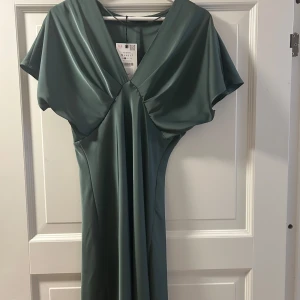 Grön klänning med v-ringning - Elegant grön klänning med v-ringad design och kortärmade ärmar. Klänningen har en mjuk och flytande passform. Passar till finare tillfällen men även andra tillfällen i sommar. Helt oanvänd från Zara och prislappen sitter kvar, slutsåld i denna färg på Zara!
