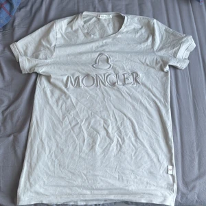 Moncler T-shirt  - Säljer en stilren vit t-shirt från Moncler med korta ärmar. T-shirten har en broderad Moncler-logga på framsidan som ger en exklusiv känsla. Perfekt för en avslappnad men ändå elegant look.