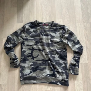 Camouflage mönstrad tröja och byxor - Säljer en set med tröja och byxor i grått och svart camouflage mönster. Tröjan har långa ärmar och byxorna har en bekväm resår med snörning i midjan. Storlek S. Noppriga på vissa ställen går säkert att få bort med noppmaskin annars bra skick! 