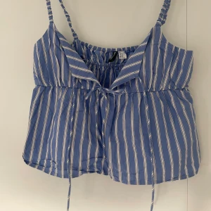 Blå- och vitrandig linnetopp med knyt - Säljer en blå linnetopp med vita ränder och smala axelband. Toppen har knyt framtill och en lätt volang vid bysten. Perfekt för varma dagar och har en luftig passform.
