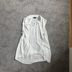 Vit blus från Amelie & Me - Säljer en stilren vit blus från Amelie & Me. Blusen har en v-ringad design med volangdetaljer vid kragen och är ärmlös. Perfekt för en elegant och luftig look. Passar bra till både jeans och kjol.