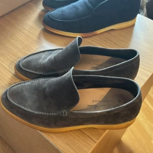 Mörkbruna summerwalks från Loro Piana - Säljer ett par stilrena mörkbruna loafers från Loro Piana, modellen Summer Walk. Skorna är i mjuk mocka med ljusbruna gummisulor och har en klassisk, rund tå. Perfekta för en avslappnad men elegant look. Är öppen för byten! Det är en liten spricka i sulan! Men funkar ändå! 