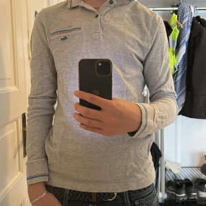 Grå långärmad pikétröja från Lacoste - Säljer en stilren grå långärmad pikétröja från Lacoste med klassisk krage och knappar. Tröjan har färgade detaljer vid ärmslut och krage samt den ikoniska krokodilloggan på bröstet. Perfekt för en clean och sportig look. Helt oanvänd, nypris kostar den runt tusenlappen