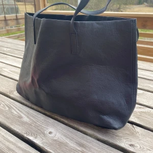 Svart axelväska från Divided - Stilren svart axelväska från Divided i slätt skinnliknande material. Väskan har enkel design. Perfekt för att bära med sig det viktigaste till vardagen. Perfekt för att ha datorn i.