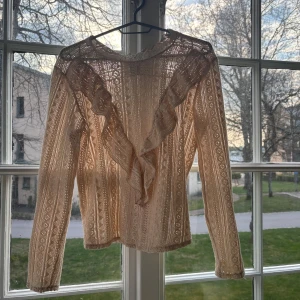 Beige spetsblus med volang från KappAhl - Säljer en superfin beige spetsblus från KappAhl i storlek XS. Blusen har långa ärmar och dekorativa volanger framtill. Perfekt för dig som gillar romantisk och detaljerad stil. Materialet är spets och känns lätt och luftigt.