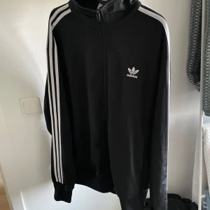 Svart träningsjacka från Adidas - Säljer en klassisk svart träningsjacka från Adidas med de ikoniska tre vita ränderna längs ärmarna. Jackan har dragkedja och Adidas-logga på bröstet. Perfekt för träning eller en avslappnad stil.