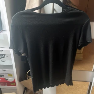 Svart ribbad t-shirt från 157 - Svart ribbad t-shirt från 157 med tight passform. T-shirten har korta ärmar och vågig kant nertill och vid ärmslut, vilket ger en snygg detalj. Perfekt för en stilren och enkel look.