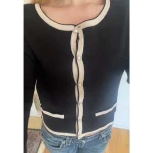 Zara: Svart kofta med pärlknappar och vit kant - Snygg svart kofta med vit kantband och dekorativa pärlknappar framtill. Koftan har två fickor fram och rund halsringning. 