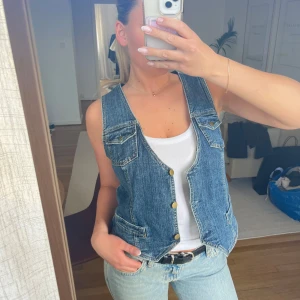 Jeansväst med knappar - Snygg jeansväst med v-ringning och knappar framtill. Västen har två fickor med lock och en klassisk blå denimfärg. 