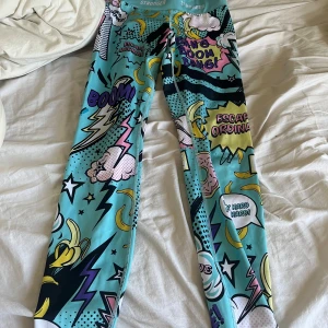 Färgglada leggings med serietidningsmönster från Stronger - Säljer ett par coola leggings från Stronger med ett färgglatt serietidningsmönster. De har en hög midja och är perfekta för att sticka ut på gymmet. Mönstret inkluderar ord som 'Boom!' och 'Escape Ordinary'. Använt dom få gånger. Köpte dom för 800 kr. 