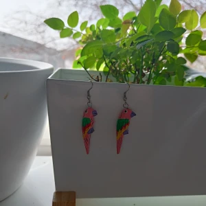 Färgglada papegojörhängen - Säljer ett par unika örhängen formade som papegojor i härliga färger. De har en mix av rosa, grön, lila och gul som ger en lekfull och somrig känsla. Perfekta för att liva upp vilken outfit som helst! Lite avskavt på undersidan av ena svansen, dock tror jag inte man kommer se det när man har dem på sig. Strass! Material: någon form av trä, skulle jag tro.