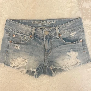 Ljusblå slitna jeansshorts från American Eagle - Säljer ett par ljusblå jeansshorts från American Eagle med slitna detaljer och fransig kant. Klassisk femficksmodell med låg midja och knappgylf. Perfekta för sommaren och en avslappnad stil.