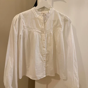 Vit blus med spetsdetalj från Nelly - Säljer en vit blus från Nelly med fina spetsdetaljer över bröstet och knappar framtill. Blusen har lång ärm med lätt puff och en avslappnad passform. Perfekt att styla med jeans eller kjol för en trendig look. Helt ny och lappen sitter kvar💕
