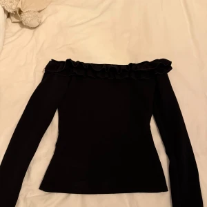 Svart offshoulder topp med volang - Säljer en svart offshoulder topp från Nelly i storlek XS. Toppen har långa ärmar och en snygg volangdetalj längs den bara axeln. Perfekt för dig som vill ha en stilren och trendig look.