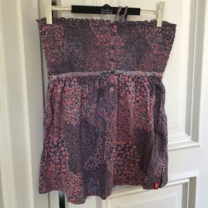 Blommig topp från  EDC - Säljer en söt blommig topp/klänning från edc i rosa och lila toner. Klänningen är axelbandslös med en smockad överdel och ett fint blommönster. Perfekt för varma dagar!