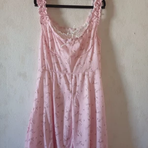 Rosa blommig klänning med spetsdetaljer - Charmig rosa klänning med blommönster och volangprydda axelband. Klänningen har en söt spetskant vid halsringningen och nederkanten. Perfekt för en romantisk look.