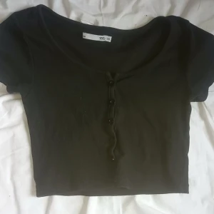 Svart croppad topp med knappar - Säljer en svart croppad topp med korta ärmar och knappar framtill. Toppen har en rundad halsringning och är tillverkad i ett ribbat material som ger en snygg passform.