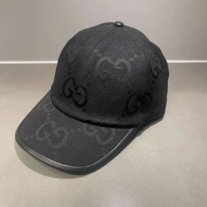Svart keps från Gucci med GG-mönster - Snygg svart keps från Gucci med ikoniskt GG-mönster ton-i-ton över hela kepsen. Justerbar rem baktill och böjd skärm. Tillverkad i slitstark bomull med detaljer i syntet. Perfekt accessoar för en trendig streetstyle-look.