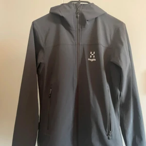 Svart Höglövs jacka - Snygg svart vindjacka från Haglöfs i dammodell, storlek S. Jackan har huva, hel dragkedja framtill och två sidofickor med dragkedja. Perfekt för blåsiga dagar. Stilren design med diskret Haglöfs-logga på bröstet.