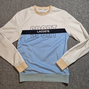 Lacoste Sport sweatshirt i blått och beige - Snygg sweatshirt från Lacoste Sport i beige och ljusblått med mörkblå detalj och stor SPORT-tryck på bröstet. Klassisk rund hals, ribbade muddar och ikoniska krokodilloggan nertill. Perfekt för en sportig och chill stil.