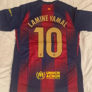 FC Barcelona Lamine Yamal #10 Nike tröja - Säljer en FC Barcelona matchtröja med Lamine Yamal och nummer 10 på ryggen. Tröjan är randig i mörkblått och rött med gula detaljer, klubbmärke och sponsorer på bröstet. Tillverkad i lätt och ventilerande material, perfekt för fotbollsälskare.
