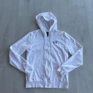 Vit zip-hoodie från EA7 Emporio Armani - Snygg vit zip-hoodie från EA7 Emporio Armani med dragkedja framtill och huva med snörning. Liten svart EA7-logga på bröstet och diskret randig detalj bak. Tillverkad i mjuk bomull, perfekt för en clean och sportig look.