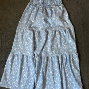 Blommig lågmidjad långkjol från Gina Tricot - Supersöt lågmidjad blå långkjol från Gina Young med små vita blommor. Kjolen har smock i midjan för extra bekväm passform och volangiga lager som ger en flowy känsla. Endast använd ett fåtal gånger och säljer den då den tyvärr är lite lång på mig💞