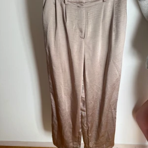 Beiga/ guldiga kostymbyxor i satinlook från Ginatricot - Snygga beiga kostymbyxor med hög midja och vida ben. Byxorna har en lyxig satinliknande finish, hällor för bälte och fickor både fram och bak. Supercoola i guld liknande färg. Tyvärr en liten fläck på baksidan. Inget man uppfattar när byxorna används! 