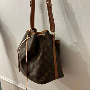 Louis Vuitton Petit Noé bag - Säljer en klassisk brun axelväska från Louis Vuitton med ikoniskt monogrammönster och justerbar axelrem i skinn. Väskan har dragskostängning med läderdetaljer och guldiga metalldelar. Remmen är utbytt nyligen från Louis Vuittons butik. Har ingen tydlig vintagelukt och är i väldigt fint skick. Dust bag från A retrotale ingår och det är därifrån väskan är inköpt. Köparen står för frakten!
