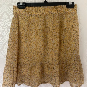 Gul blommig kjol från Vero Moda - Supersöt gul blommig kjol från Vero Moda Aware med volangkant nertill. Kjolen har ett lätt och flowy tyg, perfekt för varma dagar. Resår i midjan gör den bekväm och enkel att bära. Mönstret är småblommigt i gult och beige.