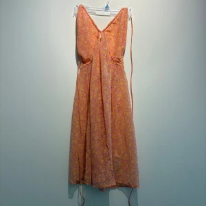 Blommig kortklänning från Bershka - Supersöt kort klänning från Bershka i orange och rosa med blommigt mönster. Klänningen har smala axelband, v-ringning och knytning fram samt smock i ryggen för extra komfort. Perfekt för varma dagar och somriga vibes!