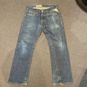 Blå jeans från Replay, midja 28/32 - Klassiska blå jeans från Replay med raka ben och snygga slitningar. Tyvärr har byxorna ett litet hål som man ser på bilden men syns inte närm an har på sig dom men bör vara enkelt att lapp igen om man vill det, därför det billiga priset.