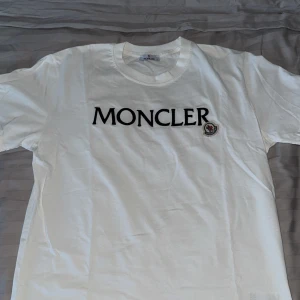 Vit Moncler t-shirt med logga - Vit t-shirt från Moncler i bomull med stor svart Moncler-logga broderad på bröstet och klassiskt rund halsringning. På bröstet finns även Monclers ikoniska patch i vitt, blått och rött. Enkel och clean design som passar till allt. Priset kan diskuteras 