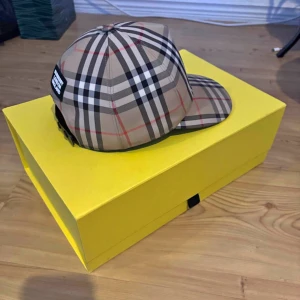 Burberry rutig keps i beige - Snygg keps från Burberry i klassiskt beige rutigt mönster med svarta, vita och röda linjer. Justerbar baktill för perfekt passform. Ikonisk streetstyle-vibe och passar perfekt till både city och chill. Kommer med originalkartong och Kvitto. 