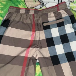 Burberry rutiga shorts med snörning - Snygga shorts från Burberry med klassiskt rutigt mönster i beige, svart, grått och rött. Shortsen har elastisk midja med svart snörning och praktiska fickor. Perfekta för sommaren och strandhäng, tillverkade i lätt polyester som torkar snabbt.