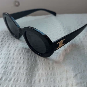 Svarta ovala solglasögon från Celine - Snygga svarta solglasögon från Celine med ovala bågar och ikonisk gulddetalj på sidorna. Glasögonen har en chunky retrostil och mörka linser.