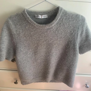 Grå stickad kortärmad tröja Zara - Mysig grå stickad tröja från Zara med korta ärmar och rund hals. Tröjan har en croppad passform och ribbade kanter vid hals och ärmslut. Perfekt att styla med höga jeans eller kjol för en chill look. Lite nopprig därav priset🥰