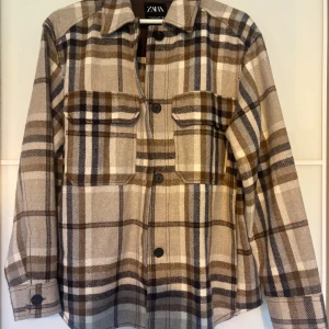 Rutig overshirt från Zara i beige och brunt - Overshirt från Zara i rutigt mönster med beige, bruna och vita toner. Skjortan har två stora bröstfickor med lock, klassisk krage och knäppning framtill. Perfekt lager-på-lager-plagg med lång ärm och avslappnad passform.