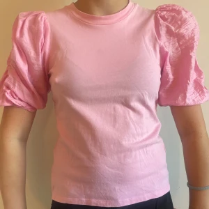 Rosa topp med puffärmar - Söt rosa topp med korta puffärmar och rund halsringning. Ärmarna har en glansig finish som ger extra volym och en cool kontrast mot den släta bomullstrikån på kroppen. Perfekt för dig som vill sticka ut lite extra med en feminin touch.