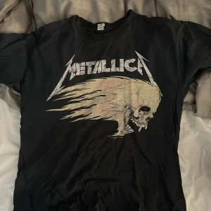 Metallica Summer Sh*t '94 t-shirt Gildan - Svart Metallica t-shirt från Gildan med stort tryck av bandets logga och en dödskalle med eldflammor på framsidan. Baksidan har turnélistan och en solformad dödskalle. Klassisk rund hals och korta ärmar, perfekt för dig som älskar rock och bandmerch.