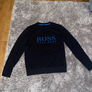 Svart tröja med blå BOSS-logga - Svart långärmad tröja från Hugo Boss med stor blå BOSS-logga på bröstet. 100% äkta köpt på Hanzens, auktoriserad återförsäljare. Tröjan har QR-kod som du kan scanna för att verifiera äktheten. Sparsamt använd och i fint skick.