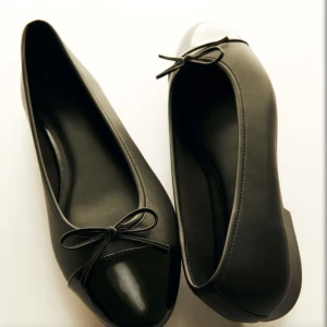 Svarta ballerinaskor med lackad tå - Stilrena svarta ballerinaskor med rund tå och lackad tådel. Skorna har en söt rosett framtill och är tillverkade i syntetmaterial. Perfekta för dig som gillar klassisk och enkel stil med en liten twist. Trendiga och slutsålda.