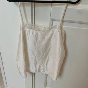 Vit spetsdetaljerad linnetopp  - Säljer en vit linnetopp med tunna axelband och fina spetsdetaljer längs överkanten. Toppen är tillverkad i glansig polyamid-nylon som ger en lyxig känsla. Perfekt att styla med jeans eller kjol för en trendig look.