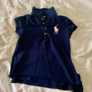 Marinblå pikétopp från Ralph Lauren - Klassisk marinblå pikétopp från Ralph Lauren med korta ärmar och rosa broderad logga på bröstet. Topp med krage och knappar framtill, tillverkad i mjuk bomull. Perfekt för en sportig och stilren look.
