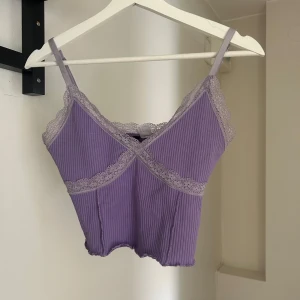 Lila ribbad linnetopp med spets - Superfin lila croppad linnetopp med tunna axelband och v-ringning. Toppen är ribbad och har dekorativa spetsdetaljer längs kanterna och under bysten. Perfekt att styla med jeans eller kjol för en trendig look.