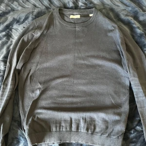 Mörkgrå tröja från Jack & Jones - En stilren mörkgrå stickad tröja från Jack & Jones med rund halsringning och ribbade muddar vid ärmslut och nederkant. Perfekt för dig som gillar en enkel och clean look. Långärmad och i mjukt bomullsmaterial. Slim för och passar M-L
