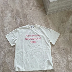 Vit Acne Studios t-shirt med tryck - Vit t-shirt från Acne Studios med rosa texttryck på bröstet där det står 'Acne Studios STOCKHOLM 1996'. Klassisk passform, rund hals och korta ärmar. Perfekt basic med en clean vibe och snygg branding.